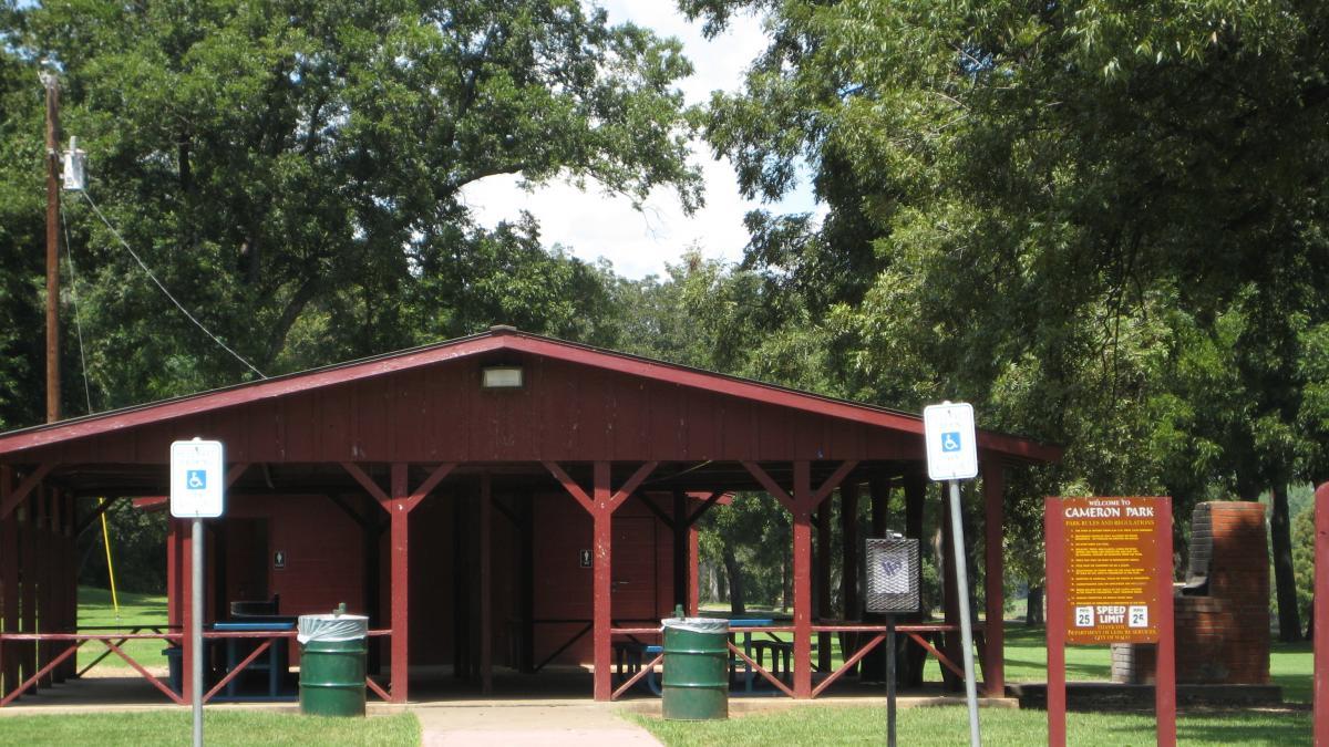 A red pavilion with a sloped roof in Cameron Park, surrounded by green grass and large trees. There are nearby trash bins and a sign welcoming visitors to the park, displaying information about park rules and a speed limit. Cameron Park mountain bike trail.