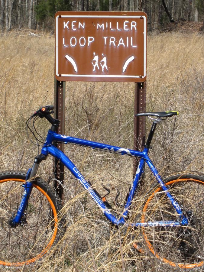 A blue mountain bike with orange rims is leaning against a rust-colored sign that reads "KEN MILLER LOOP TRAIL," surrounded by tall grass and trees in the background. South Rockdale Community Park mountain bike trail.