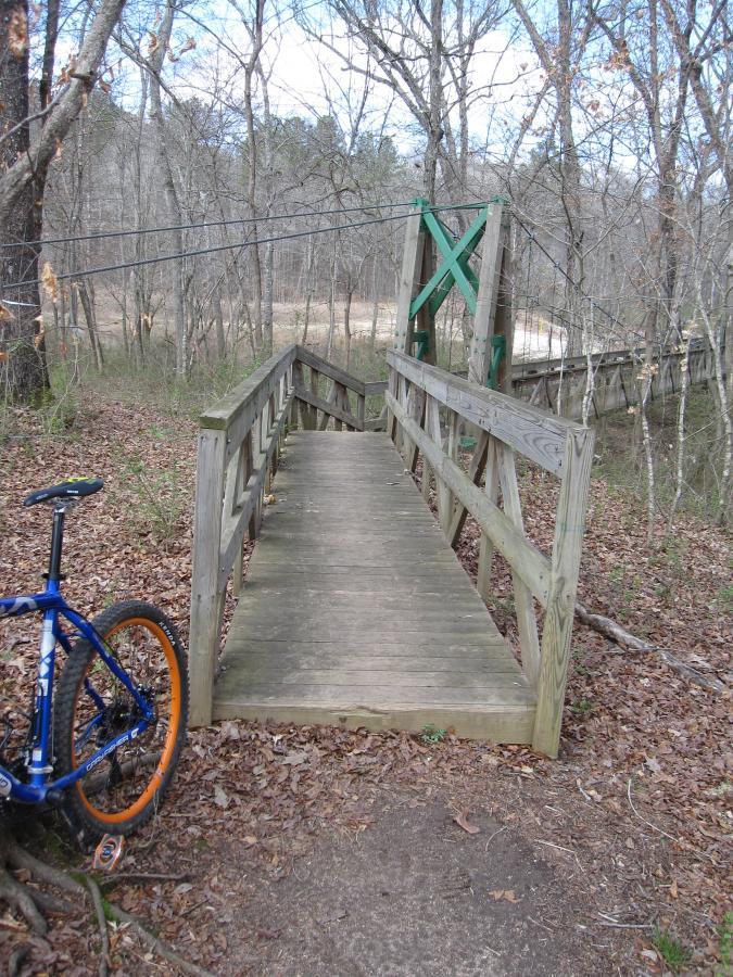 A narrow wooden bridge leading over a small stream, surrounded by trees in a natural setting. A blue mountain bike with orange wheels rests on the ground to the left of the bridge, and fallen leaves cover the ground. The sky is partly cloudy, suggesting a cool day. South Rockdale Community Park mountain bike trail.
