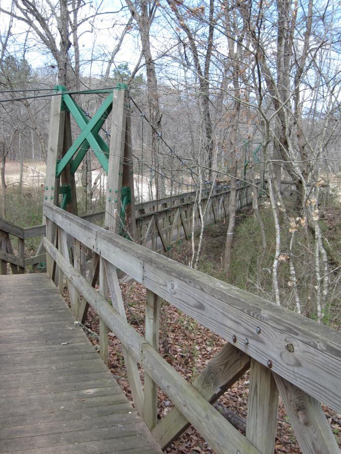 A wooden suspension bridge surrounded by bare trees and a forested area, with green support beams visible at each end. The bridge extends across a ravine, with a path leading toward its wooden planking. The scene is set in a natural environment with fallen leaves on the ground. South Rockdale Community Park mountain bike trail.