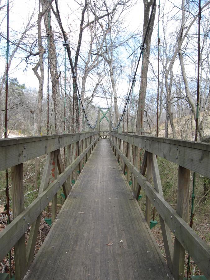 A wooden suspension bridge extending through a wooded area, with trees on either side and a clear blue sky above. The bridge features vertical support cables and a smooth wooden planked walkway, leading toward an open area in the distance. South Rockdale Community Park mountain bike trail.
