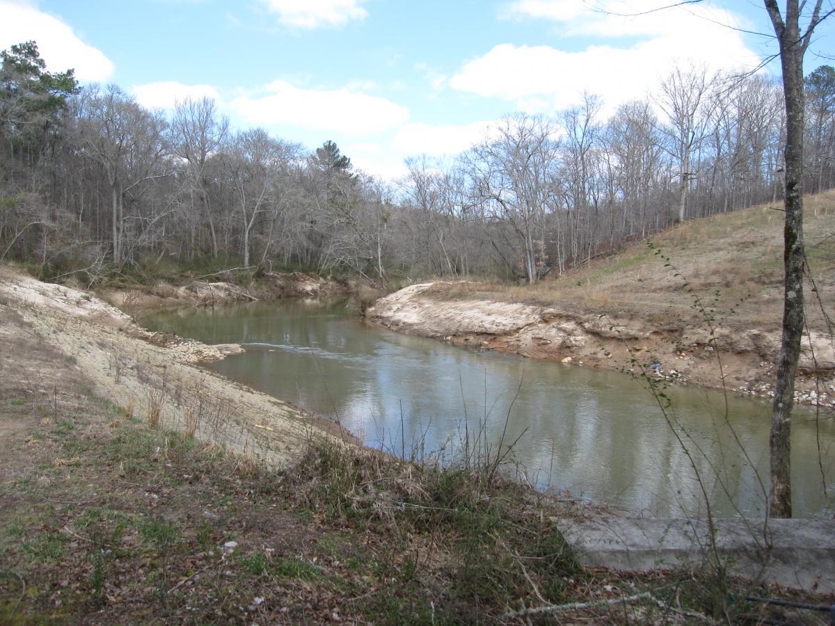 A tranquil river scene with gently flowing water surrounded by bare trees and hilly banks. The landscape features a mix of sandy and grassy areas along the riverbank, under a partly cloudy sky. South Rockdale Community Park mountain bike trail.