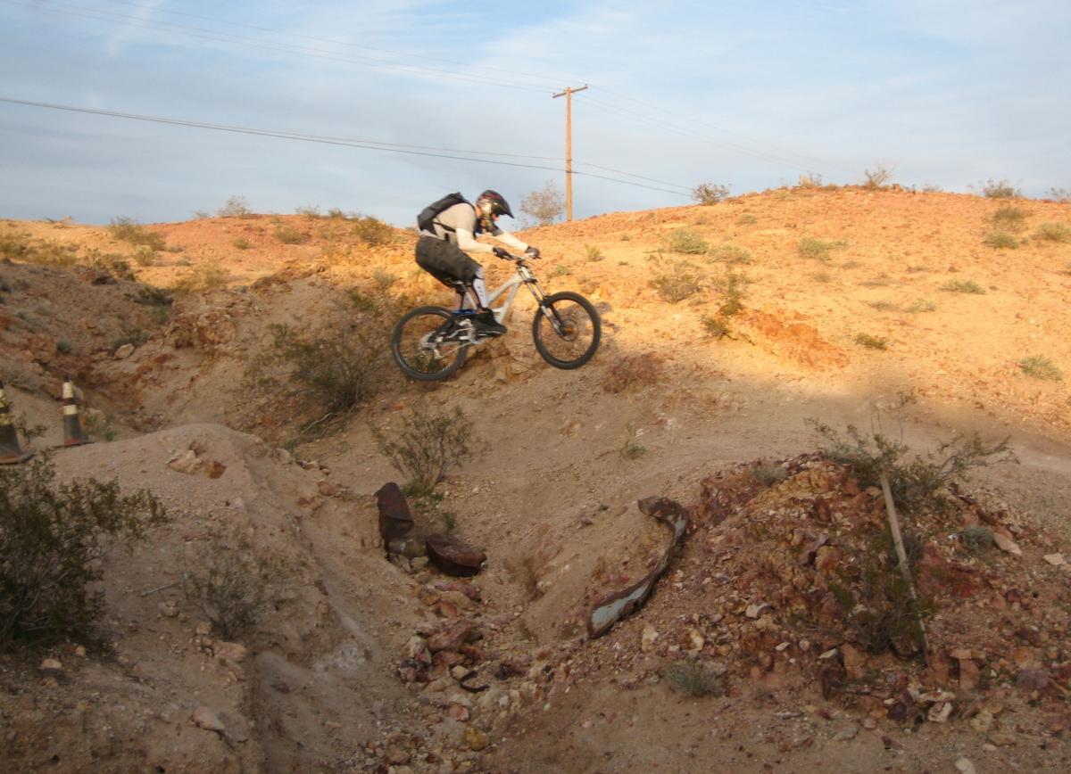 A mountain biker performing a jump over a small dirt mound in a desert landscape, surrounded by rocky terrain and sparse vegetation. The sky is partly cloudy, and power lines are visible in the background. Bootleg Canyon mountain bike trail.