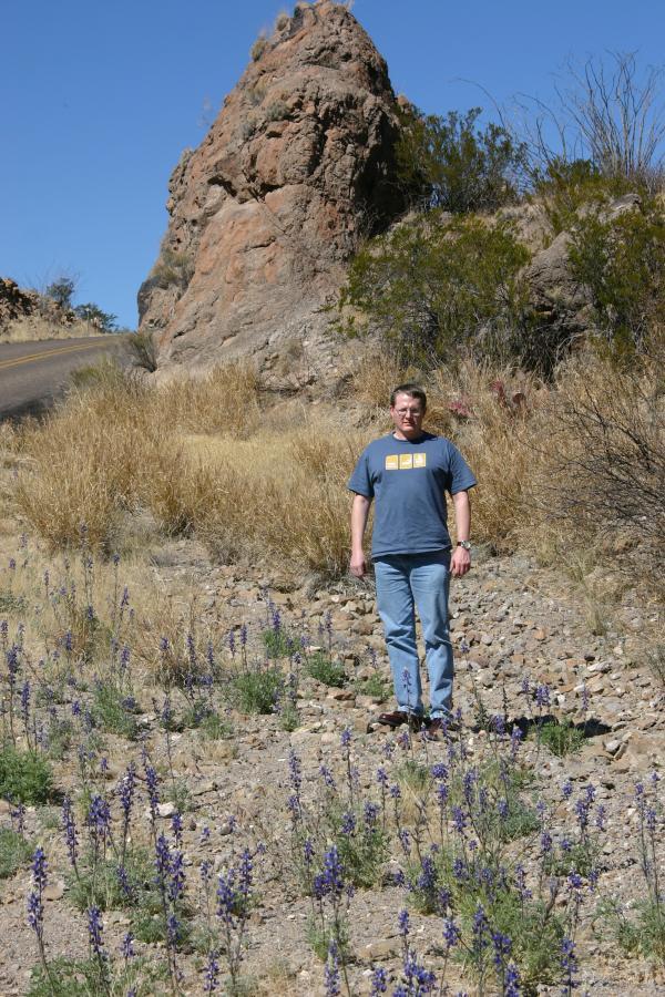A person standing on rocky terrain surrounded by purple wildflowers, with a large rock formation and a dry landscape in the background. The sky is clear and blue. Lajitas Airport Trail Head mountain bike trail.