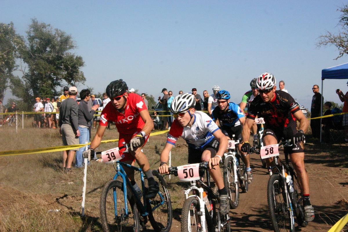 A group of mountain bikers racing in a competition, with several cyclists in colorful jerseys and helmets navigating a dirt path. Spectators are visible in the background, watching the event. The scene captures the dynamic and competitive spirit of the race. Mellow Johnnies mountain bike trail.