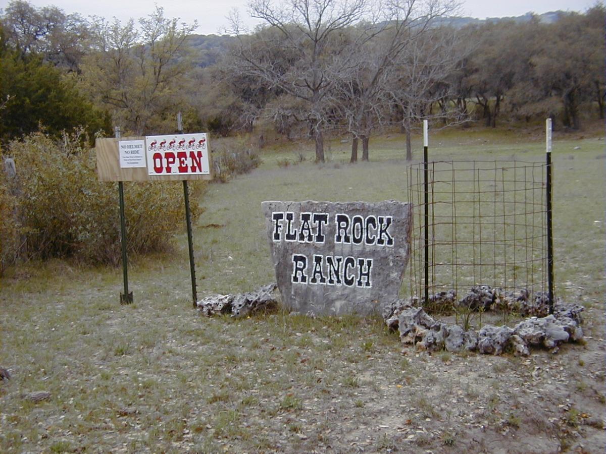 Signage at the entrance of Flat Rock Ranch, featuring a large stone marker with the ranch name, alongside a wooden sign indicating "OPEN" and safety rules stating "NO HELMET, NO RIDE." The surrounding area is grassy with sparse trees and a rural landscape. Flat Rock Ranch mountain bike trail.