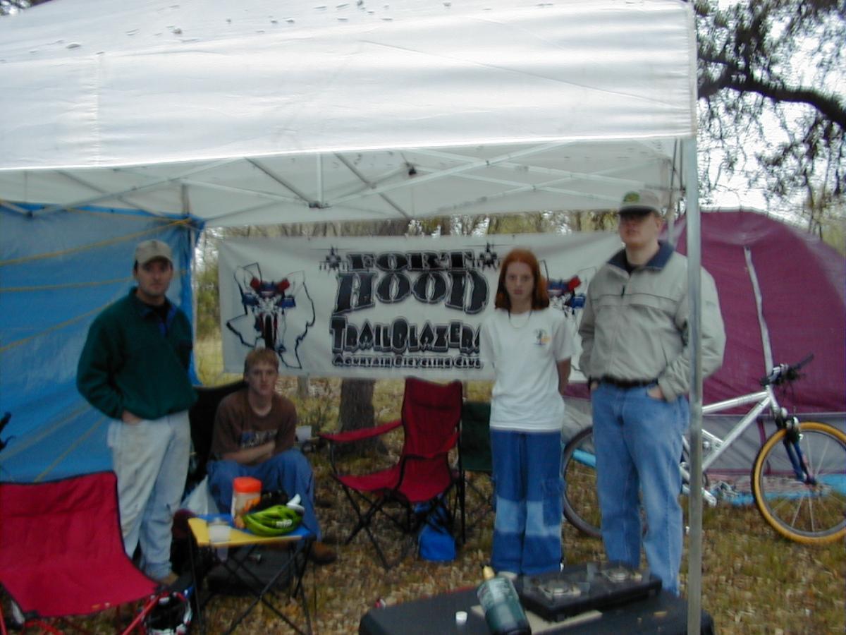 Group of four individuals standing under a camping canopy with a banner that reads "Four Hood Trailblazers." Two are seated on folding chairs, while the others stand beside them. In the background, there's a tent and a bicycle. The scene is set in a wooded area with leaves on the ground. Flat Rock Ranch mountain bike trail.