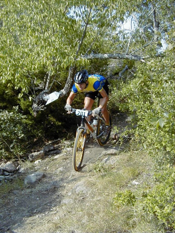 A mountain biker in a blue and yellow jersey rides down a rocky trail. The cyclist is focused and appears to be navigating through a wooded area with greenery and trees surrounding the path. The bike has yellow wheels, and a race number is visible on the front of the bike. Kelly Creek mountain bike trail.