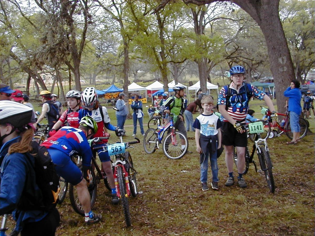 A group of cyclists gathers in a park for a mountain biking event. Several riders in colorful jerseys prepare their bikes, while a boy stands beside an older cyclist wearing an American-themed shirt. Tents and trees are visible in the background, creating a lively outdoor atmosphere. Flat Rock Ranch mountain bike trail.
