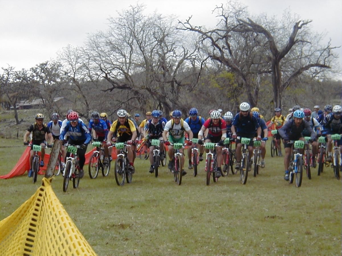 A group of mountain bikers in colorful jerseys and helmets race on a grassy field, with trees in the background. They are positioned at the starting line, ready to begin a competition, with safety barriers visible on either side. The scene captures the excitement and energy of the event. Flat Rock Ranch mountain bike trail.