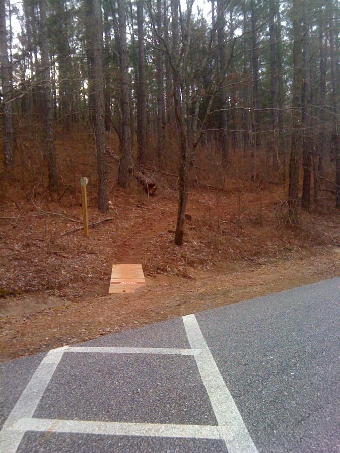 A dirt path leading into a wooded area, with a wooden ramp at the entrance. A trail marker stands near the path on the left, while a paved road with a parking area is visible in the foreground. The scene is surrounded by tall trees and dry leaves, indicating a natural environment. Lakeside Trail mountain bike trail.