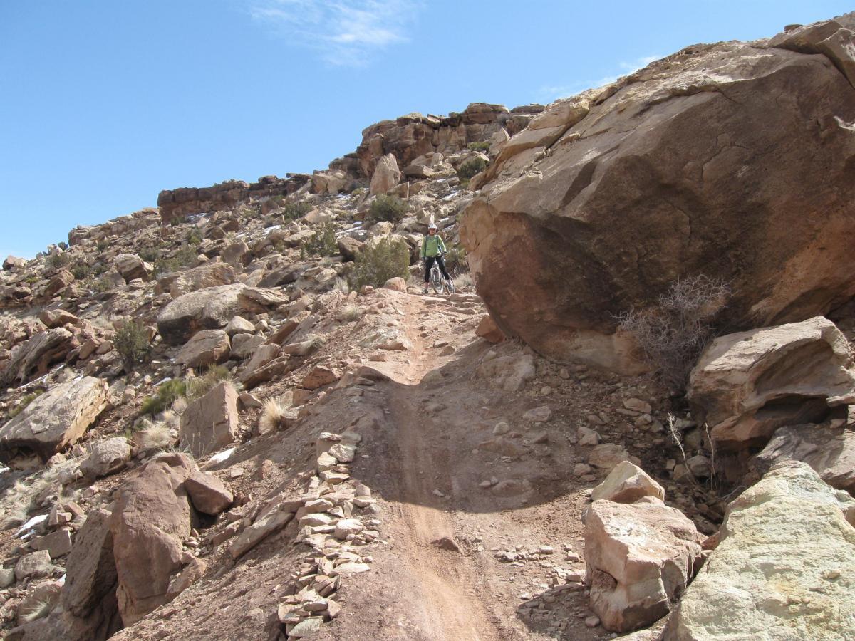 A rocky mountain trail with a cyclist navigating the uneven terrain, surrounded by boulders and sparse vegetation under a clear blue sky. Sovereign Single Track mountain bike trail.
