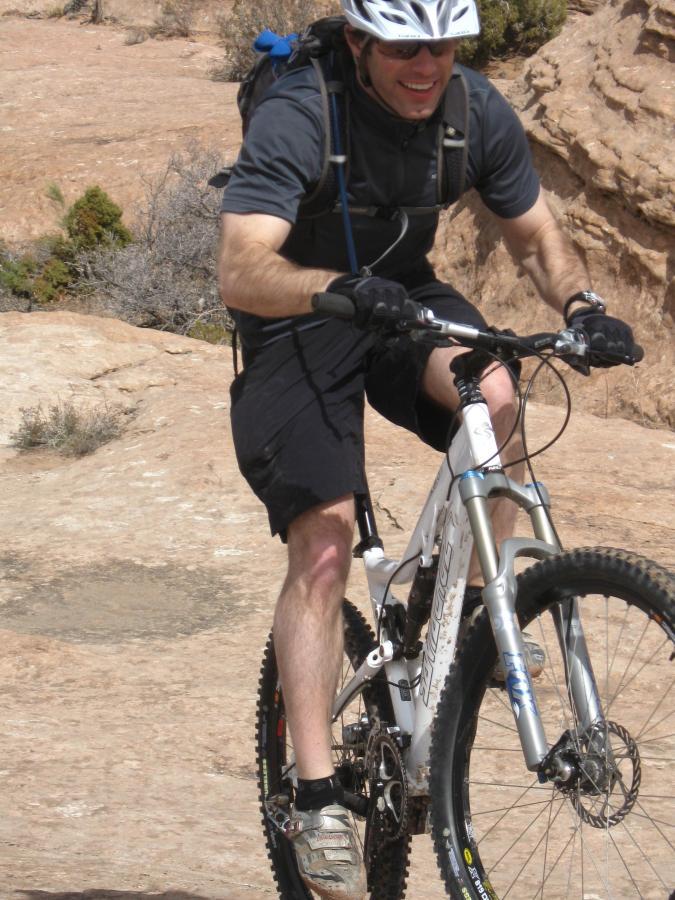 A mountain biker wearing a helmet and sunglasses is riding uphill on a rocky terrain. He is smiling and dressed in a black short-sleeve shirt and shorts, with a backpack secured to his back. The surrounding landscape features dry brush and rocky outcrops. Sovereign Single Track mountain bike trail.
