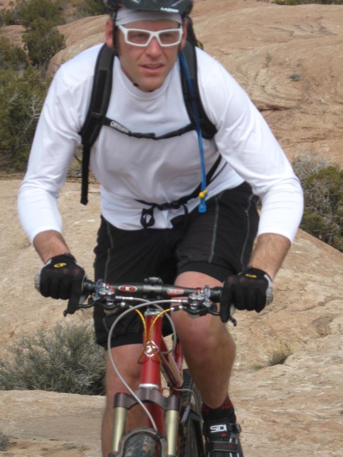A person wearing a white long-sleeve shirt, black shorts, and gloves is riding a mountain bike on rocky terrain. They have a helmet and sunglasses on, and the background features sparse vegetation and rocky landscape. Sovereign Single Track mountain bike trail.