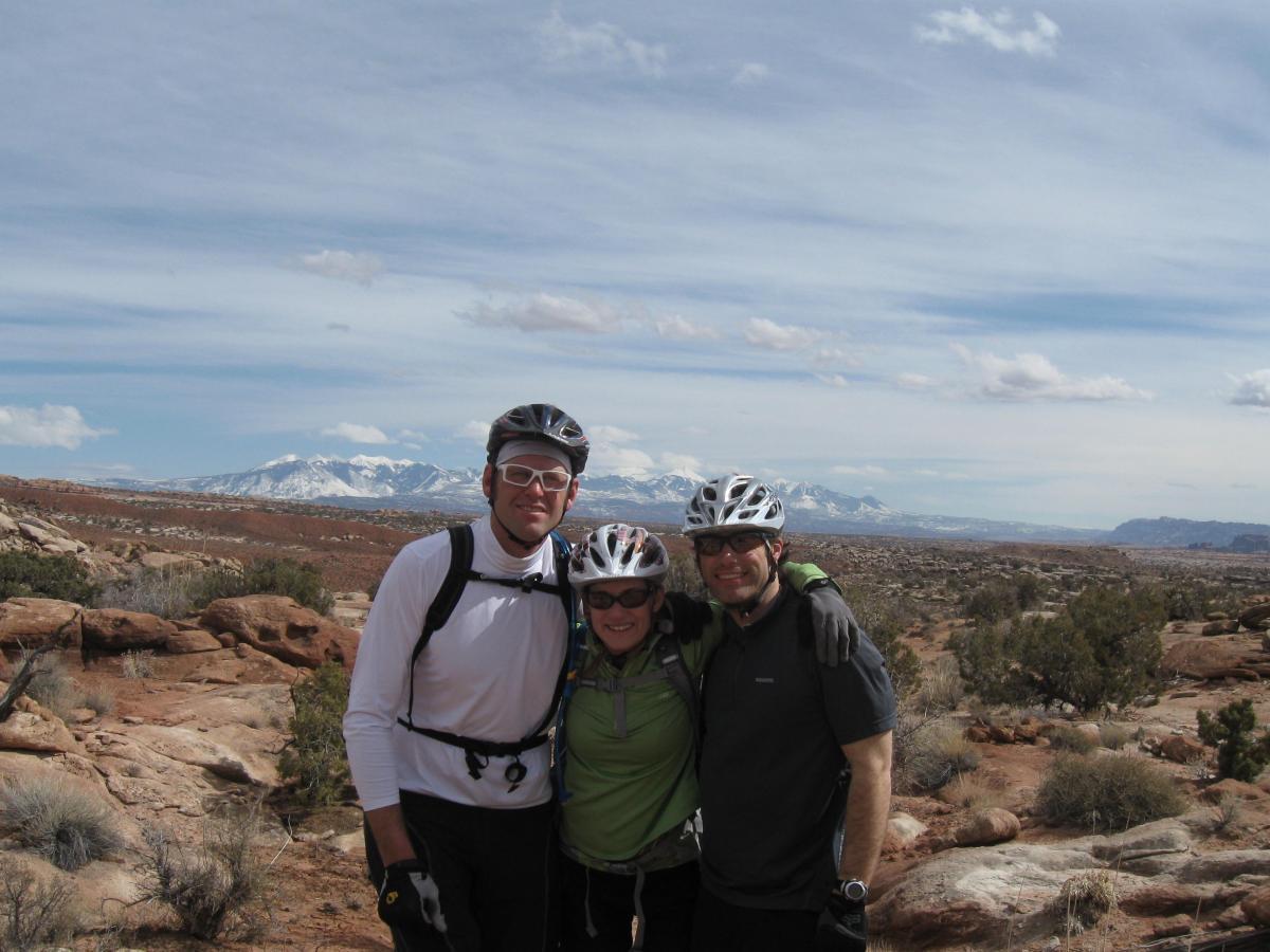 Three cyclists pose for a photo outdoors, surrounded by rocky terrain and distant snow-capped mountains. The group wears cycling gear and helmets, with the landscape featuring a mix of shrubs and boulders under a partly cloudy sky. Sovereign Single Track mountain bike trail.