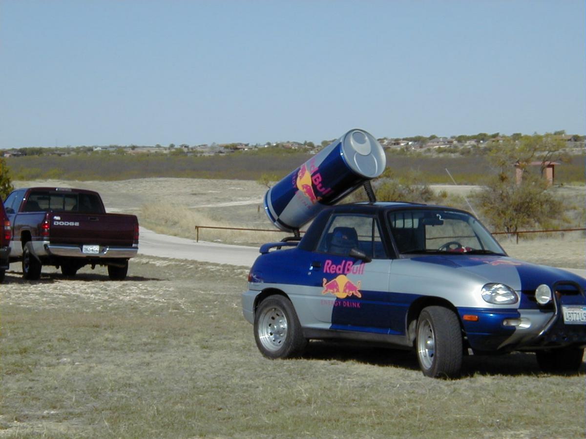 A small car decorated with Red Bull branding, featuring a large canister mounted on the roof, parked on an open terrain with sparse vegetation. In the background, another truck is also parked on a dirt road under a clear sky. San Angelo Trail mountain bike trail.