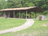 A shaded picnic pavilion with a wooden roof and several picnic tables underneath, surrounded by lush green grass and trees. A paved walkway leads up to the pavilion, and there is a trash bin nearby. South Rockdale Community Park mountain bike trail.