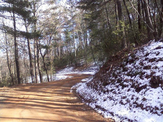 A winding dirt road surrounded by trees, with patches of snow along the edges and on the ground. The scene captures a peaceful, natural environment with dappled sunlight filtering through the foliage. Bull / Jake Mountain mountain bike trail.