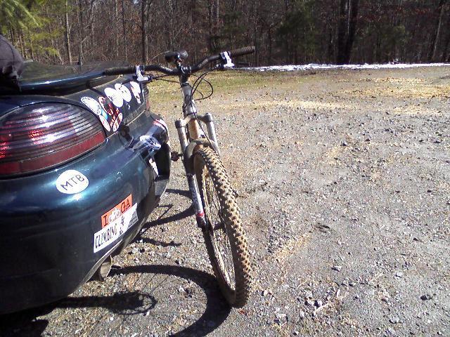 A close-up view of a mountain bike leaning against the rear of a parked car, adorned with various stickers. The background features a gravel area with sparse vegetation and trees, suggesting a natural outdoor setting. Jake to Bull Mountain Connecter mountain bike trail.
