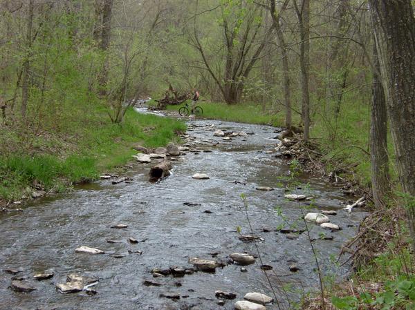 A tranquil stream flows through a lush green landscape, with smooth stones scattered along its banks. In the background, a person on a bicycle can be seen riding along a path beside the water, surrounded by trees in spring foliage. The scene captures the beauty of nature and outdoor activity. Baird Creek mountain bike trail.