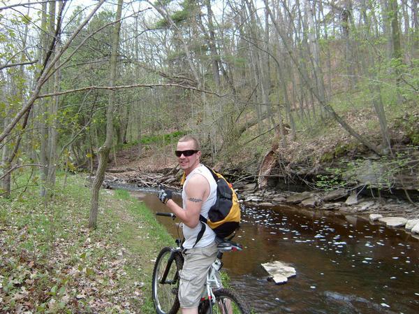 A person wearing sunglasses and a sleeveless shirt stands next to a bicycle, smiling and giving a thumbs up. They are by a creek surrounded by a lush forest with green foliage and rocky terrain. The scene captures a serene outdoor setting during the spring. Baird Creek mountain bike trail.