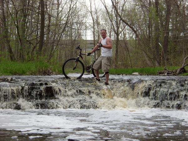 A person stands on a rocky ledge in a stream, posing with a mountain bike. The individual, wearing a sleeveless shirt and cargo shorts, smiles and gives a thumbs-up. Water cascades over the rocks behind them, surrounded by trees and greenery in a natural setting. Baird Creek mountain bike trail.