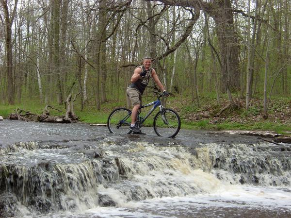 A person on a mountain bike, smiling and posing with one hand raised, stands on a rocky surface where water cascades over a small waterfall. The surrounding area features lush green trees and foliage, indicating a natural, outdoor setting during the day. Baird Creek mountain bike trail.
