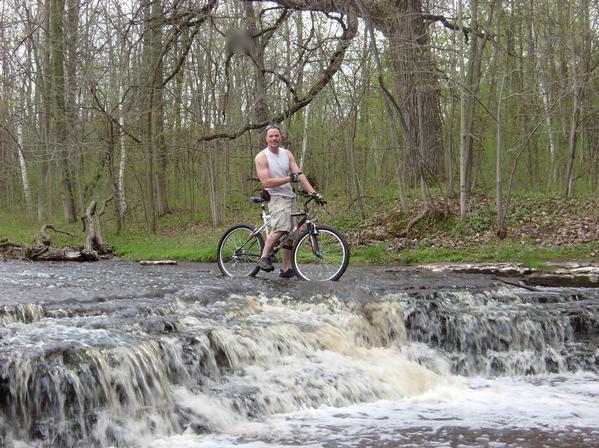 A person standing on a rocky ledge in front of a small waterfall, with a mountain bike beside them. The surrounding area features lush green trees and a flowing stream, suggesting a natural outdoor setting. The individual is wearing a sleeveless shirt and shorts, smiling at the camera. Baird Creek mountain bike trail.