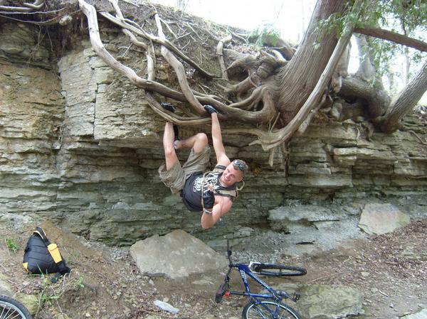 A person hanging from tree roots on a rocky cliff, wearing a tank top and gloves, with a mountain bike resting on the ground nearby. The scene is surrounded by trees and natural vegetation. Baird Creek mountain bike trail.