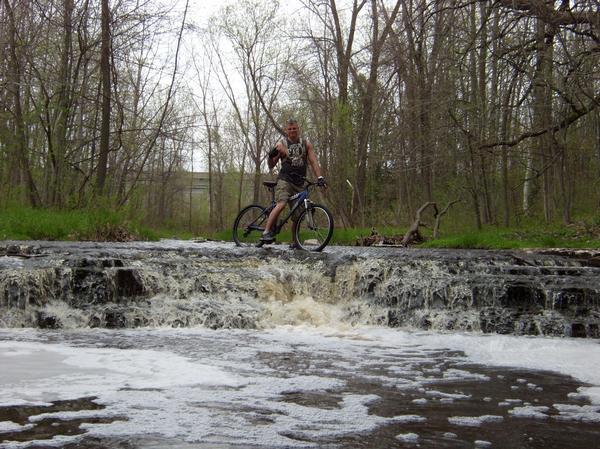 A person standing on a bicycle at the edge of a small waterfall in a wooded area, surrounded by trees and greenery. The water is flowing over rocks, creating foam at the base, while the individual poses with a raised hand. Baird Creek mountain bike trail.
