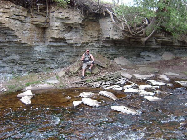 A person sitting on a rock next to a shallow stream, surrounded by a natural landscape featuring a rocky cliff and greenery. The water is gently flowing over smooth stones, and trees can be seen in the background. Baird Creek mountain bike trail.