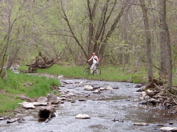 A person riding a bicycle along a grassy path beside a small stream, surrounded by trees and greenery. The scene captures a tranquil outdoor setting during springtime, with rocks scattered in the water and signs of new foliage. Baird Creek mountain bike trail.