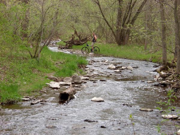 A tranquil scene featuring a winding creek surrounded by lush green trees and foliage. A person stands next to their bicycle in the shallow water, while smooth stones are scattered along the creek bed. The atmosphere is peaceful, showcasing the beauty of nature in springtime. Baird Creek mountain bike trail.