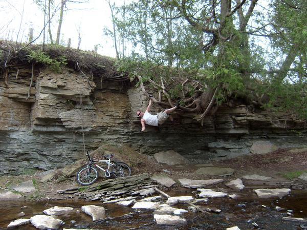 A person is hanging from the roots of a tree extending from a rocky cliff beside a stream, while a bicycle leans against the rocks nearby. The scene is set in a natural outdoor environment with greenery and stone formations. Baird Creek mountain bike trail.