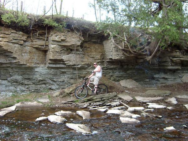 A person standing next to a bicycle beside a shallow stream, with a rocky cliff and trees in the background. The individual is wearing sunglasses and casual clothing, enjoying the natural outdoor environment. Baird Creek mountain bike trail.