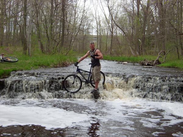 A man standing on a waterfall with his mountain bike beside him, surrounded by a forested area. The scene depicts flowing water with foam and greenery in the background. Baird Creek mountain bike trail.