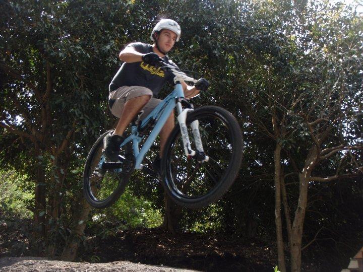 A cyclist in a helmet jumps off a dirt ramp on a blue mountain bike, with trees in the background and the bike's front wheel lifted off the ground, capturing a moment of aerial maneuver. Markham Park mountain bike trail.