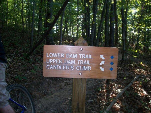 Wooden trail sign post in a forest, indicating directions for three trails: "Lower Dam Trail," "Upper Dam Trail," and "Candler's Climb," with corresponding arrows and icons. Liberty Mountain mountain bike trail.