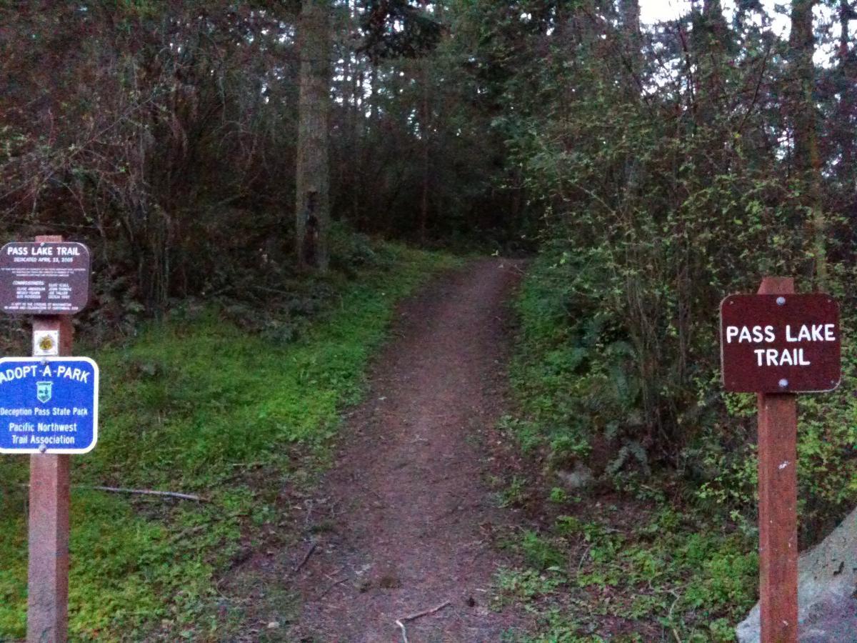 Image of a hiking trail entrance at Deception Pass State Park. Two signs are visible: one indicating "Pass Lake Trail," and another labeled "Adopt-A-Park." The trail is surrounded by dense foliage and trees, with a natural dirt path leading into the forest. The scene is set in a serene outdoor environment, suggesting a peaceful hiking opportunity. Pass Lake Trail mountain bike trail.