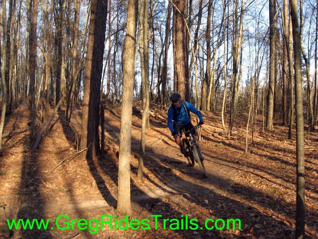 A mountain biker in a blue jacket navigates a winding trail through a wooded area with bare trees and fallen leaves. The sunlight filters through the branches, casting shadows on the path. Gainesville College mountain bike trail.