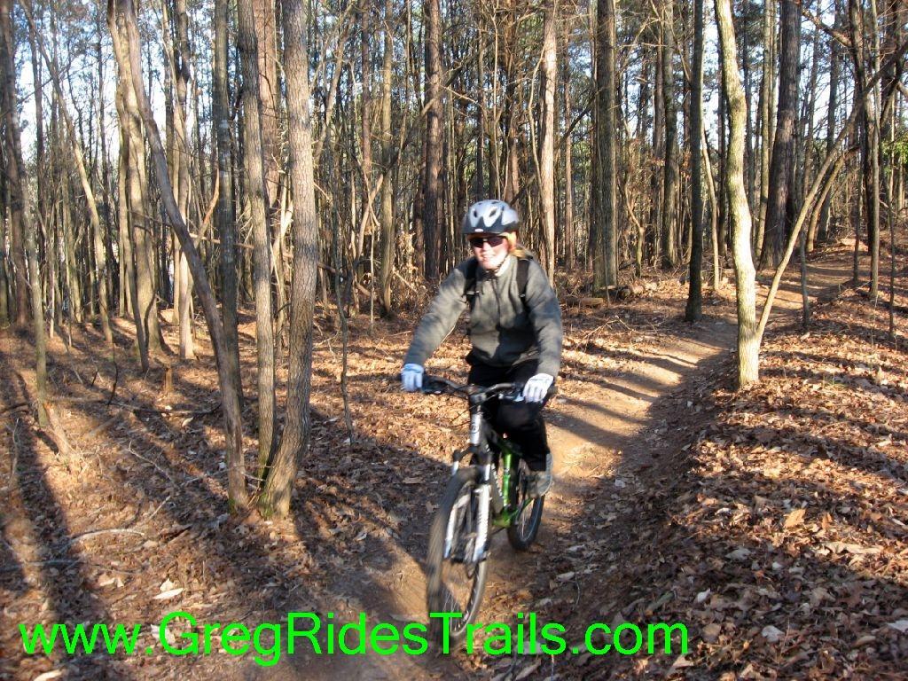 A person riding a mountain bike on a forest trail surrounded by tall trees, with fallen leaves covering the ground. The cyclist is wearing a helmet and gloves, and is smiling while navigating the winding path through the woods. Gainesville College mountain bike trail.