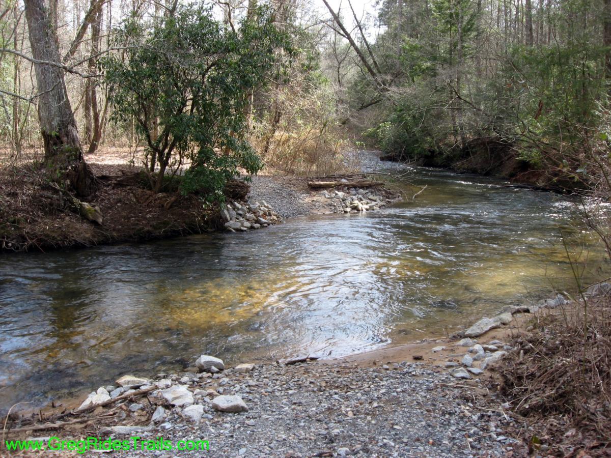 A tranquil creek flows through a wooded area, with smooth stones lining the water's edge. Lush greenery and trees border the banks, while sunlight filters through the branches, creating dappled patterns on the water's surface. Jake Mountain Trails mountain bike trail.