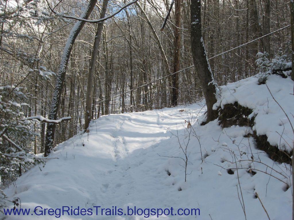 A snowy forest trail winding through a dense woods, with snow-covered trees lining the path. The ground is blanketed in fresh snow, and a slight incline is visible in the distance. Fontana Village mountain bike trail.