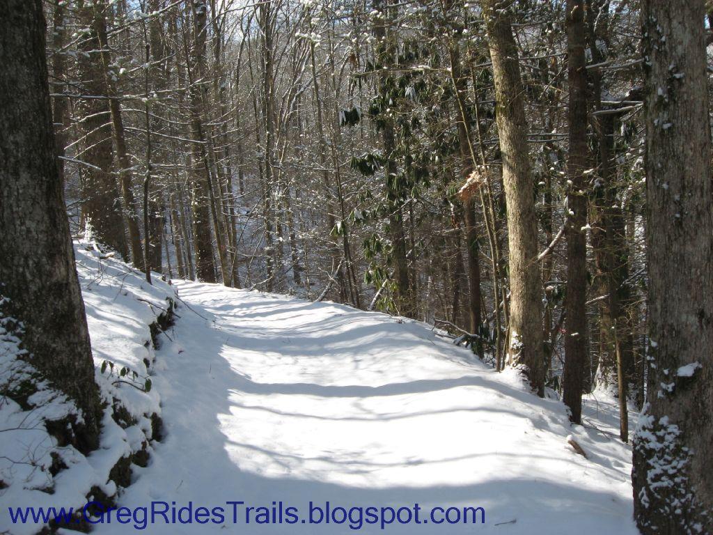 A snowy trail winding through a forest, with sunlight filtering through the trees. The path is lined with snow-covered ground and partially snow-dusted trees, creating a serene winter landscape. Fontana Village mountain bike trail.
