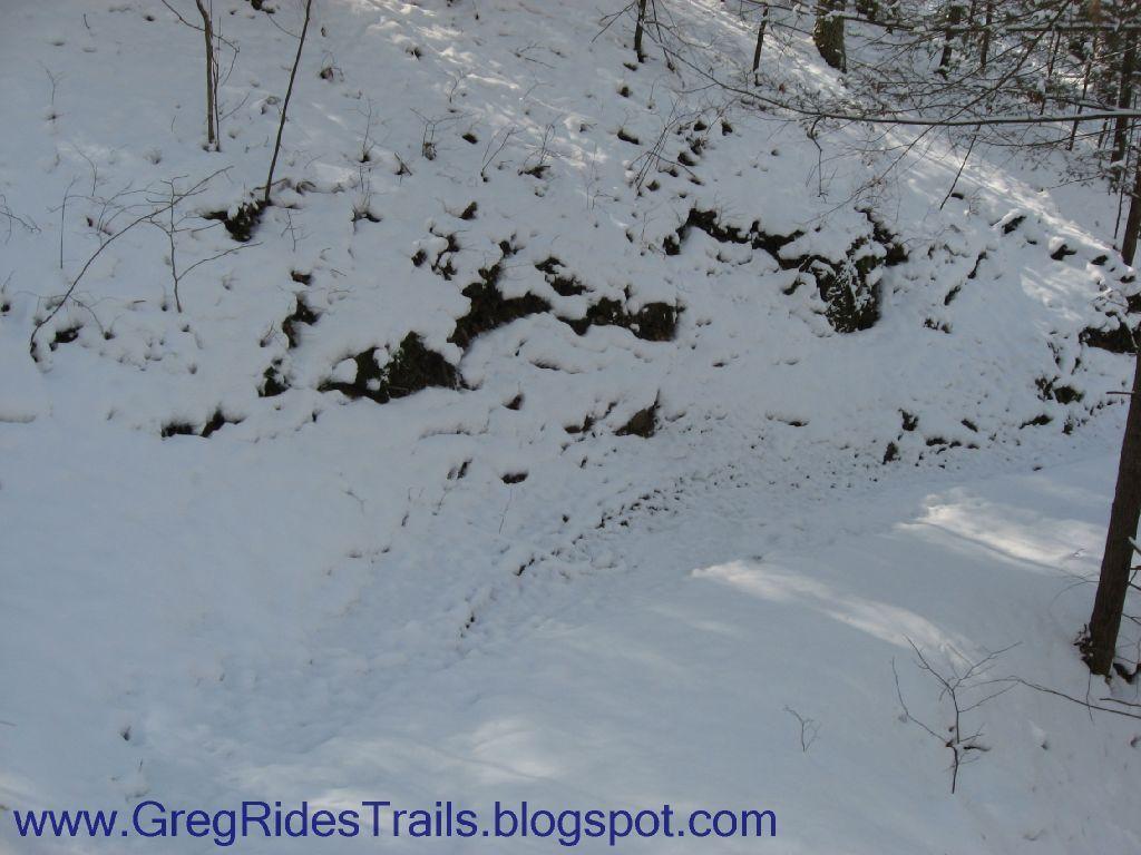 Snow-covered ground along a rocky slope in a forested area, with trees partially visible in the background. The scene conveys a tranquil winter atmosphere. Fontana Village mountain bike trail.