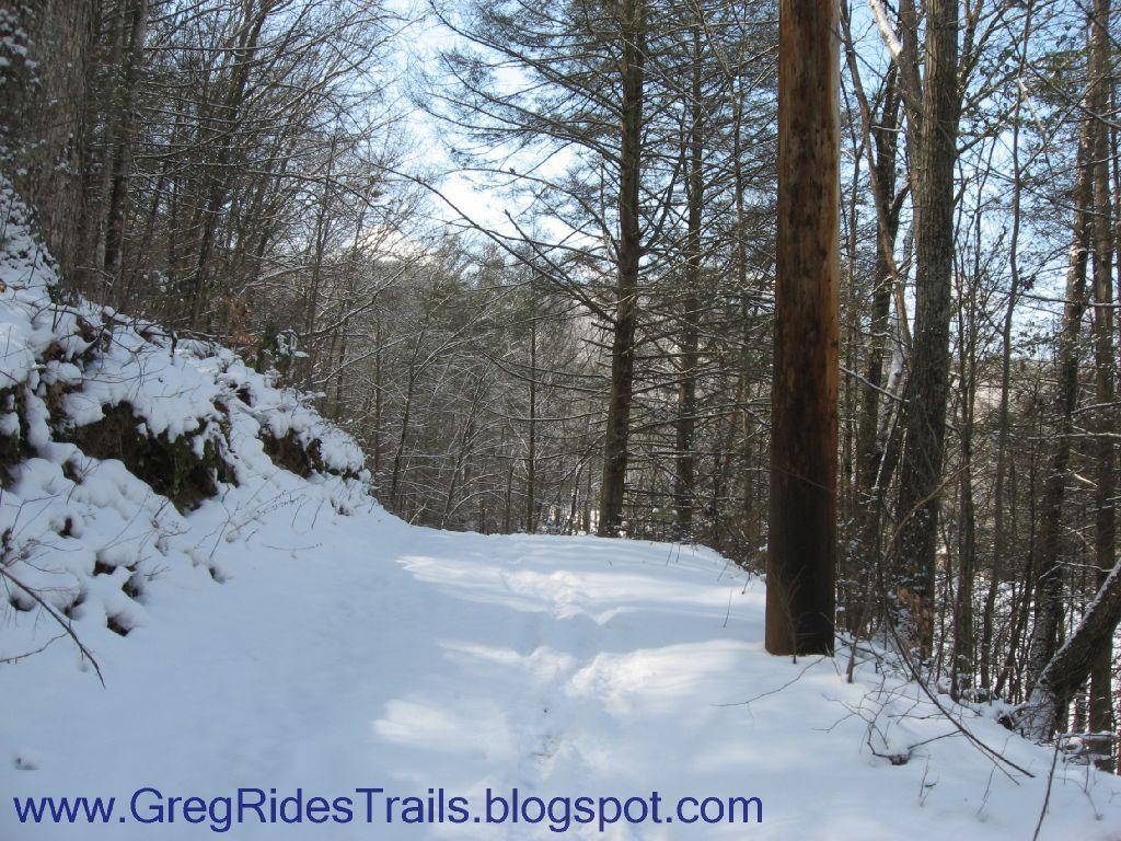 A snowy trail winding through a forest, lined with tall trees. The path is partly covered in fresh snow, indicating recent snowfall. A wooden utility pole stands next to the trail, blending into the natural surroundings. The scene conveys a peaceful winter landscape. Fontana Village mountain bike trail.