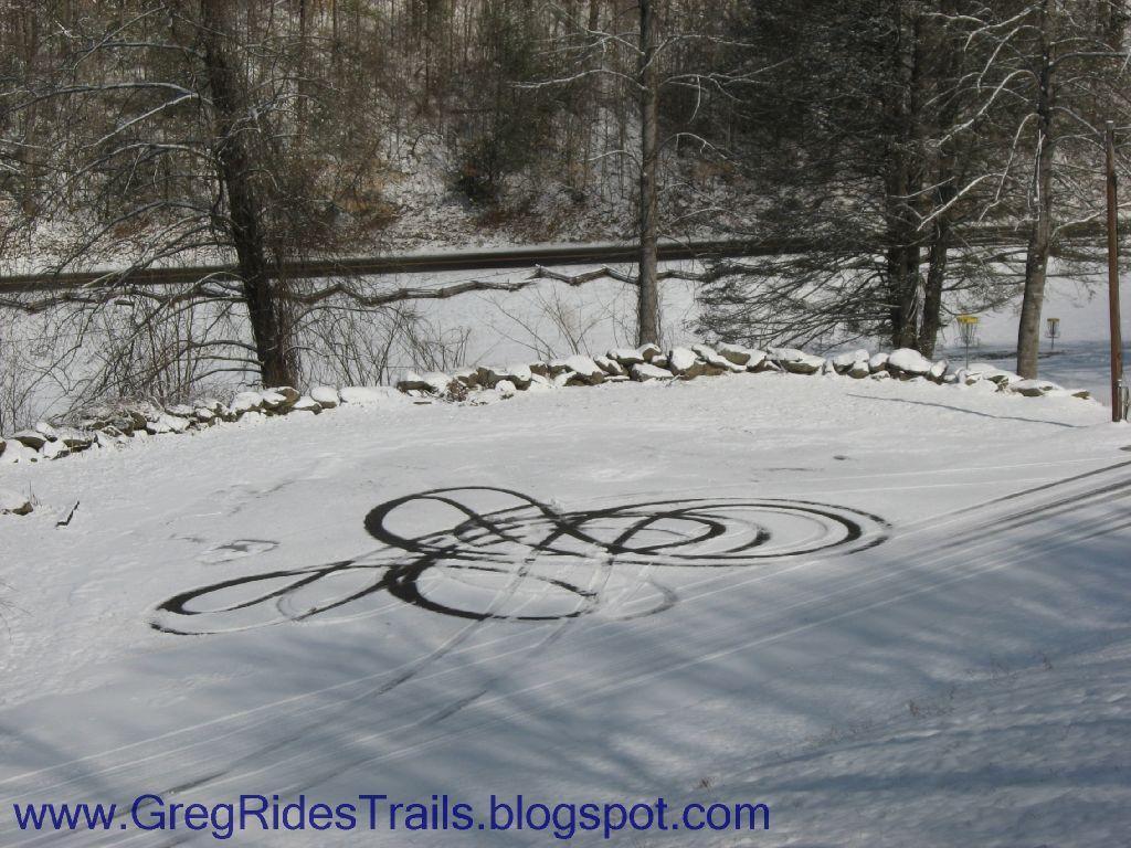 A snowy landscape featuring tire tracks that create swirling patterns on the white snow. Trees line the background, and a road is visible in the distance. The scene captures a winter day with clear skies and a serene atmosphere. Fontana Village mountain bike trail.