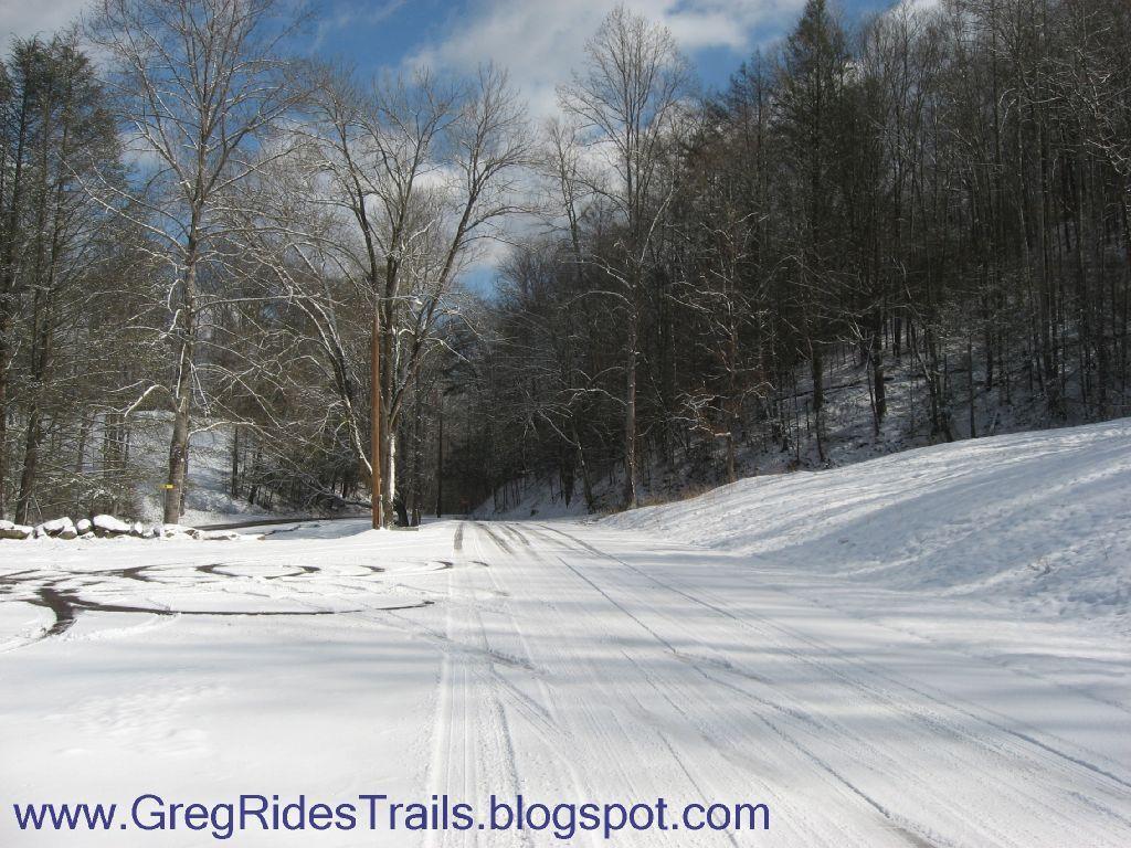 A snow-covered road lined with bare trees and a sloping hillside in the background. The scene is brightened by a partly cloudy sky, showcasing a tranquil winter landscape. Tire tracks are visible in the snow, indicating recent travel. Fontana Village mountain bike trail.