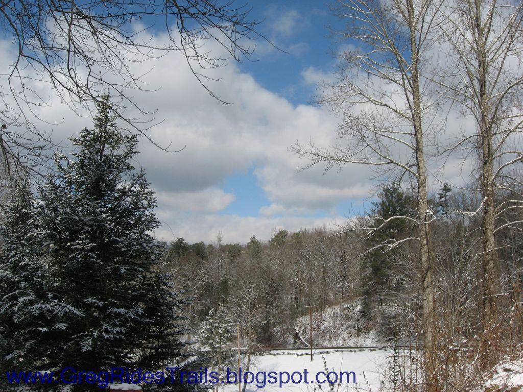 A serene winter landscape featuring snow-covered trees and a cloudy sky. In the background, a frozen body of water is partially visible, and the scene is framed by bare branches, suggesting a tranquil, chilly atmosphere. Fontana Village mountain bike trail.
