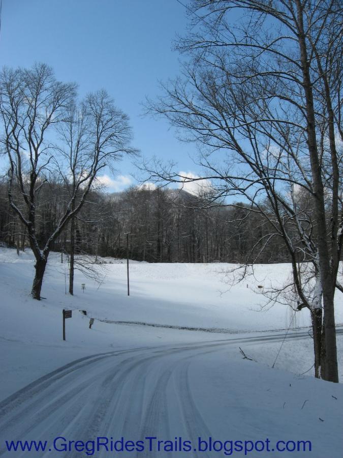A snowy landscape featuring a winding road surrounded by bare trees under a clear blue sky. The scene is tranquil, with a fresh layer of snow covering the ground, creating a serene winter atmosphere. Fontana Village mountain bike trail.